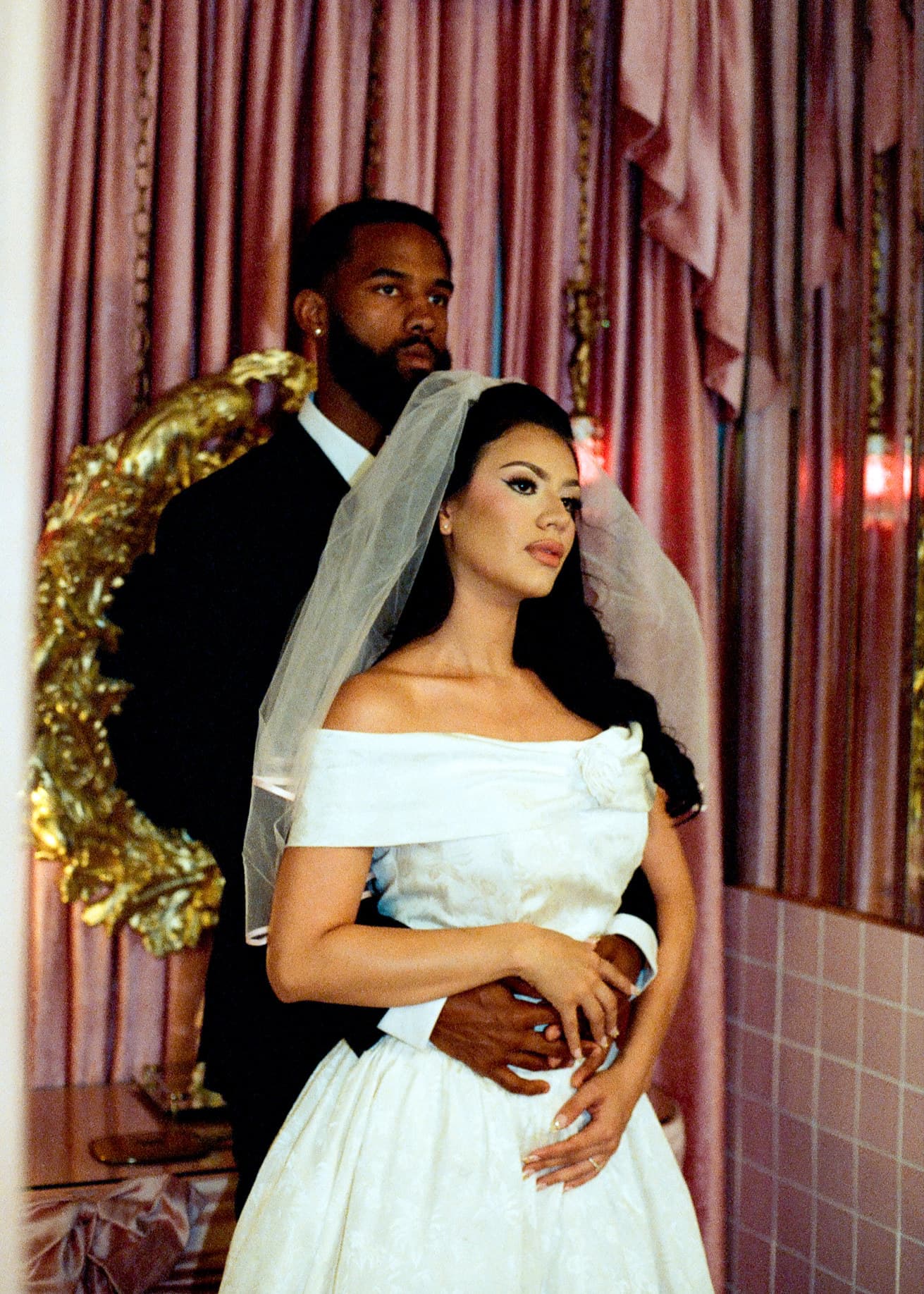 A groom in a black suit holds his bride in an off-shoulder gown and veil inside a pink-curtained chapel